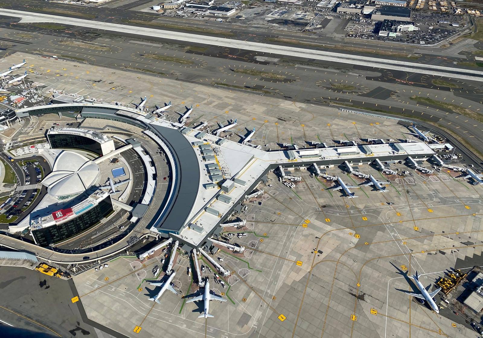 An aerial view of JFK Terminal 5 with aircraft parked at the stands around it, and the winged roof of the TWA Flight Center and the curved TWA Hotel wings in front of it on the landside